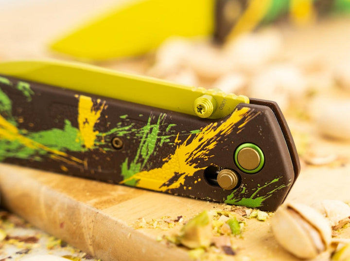 Close-up of the Böker Plus Kihon Dubai Chocolate yellow blade and splatter-pattern handle on a wooden board with pistachios