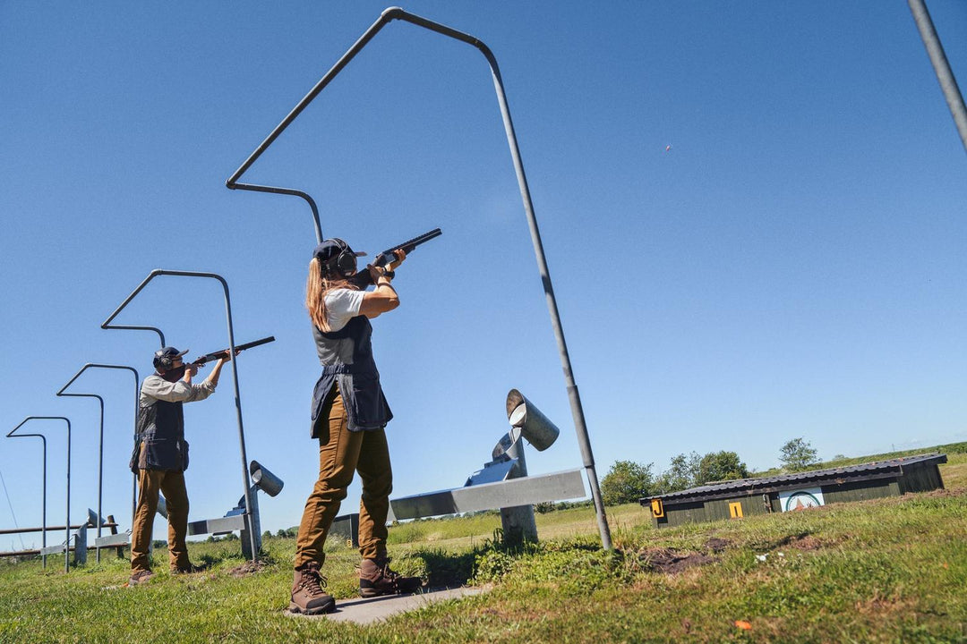 Lifestyle shot: wide view of a skeet range under blue sky, two clay shooters in Seeland shooting waistcoats at the stand