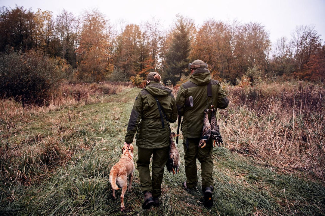 Lifestyle shot on autumn marshland: two people in olive Seeland kit walking away through long grass with a working dog at heel.