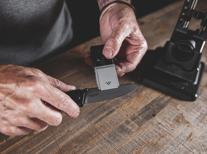 Lifestyle shot of a hand placing a folding knife into the Work Sharp Precision Adjust sharpener clamp on a wooden bench