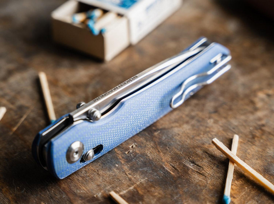 Blue Böker Magnum Shango folding knife on a wooden table beside matches and matchbox, lifestyle shot