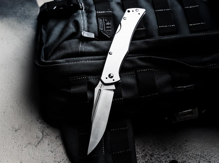 Böker Plus MEK steel folding knife open on a black tactical backpack, lifestyle shot