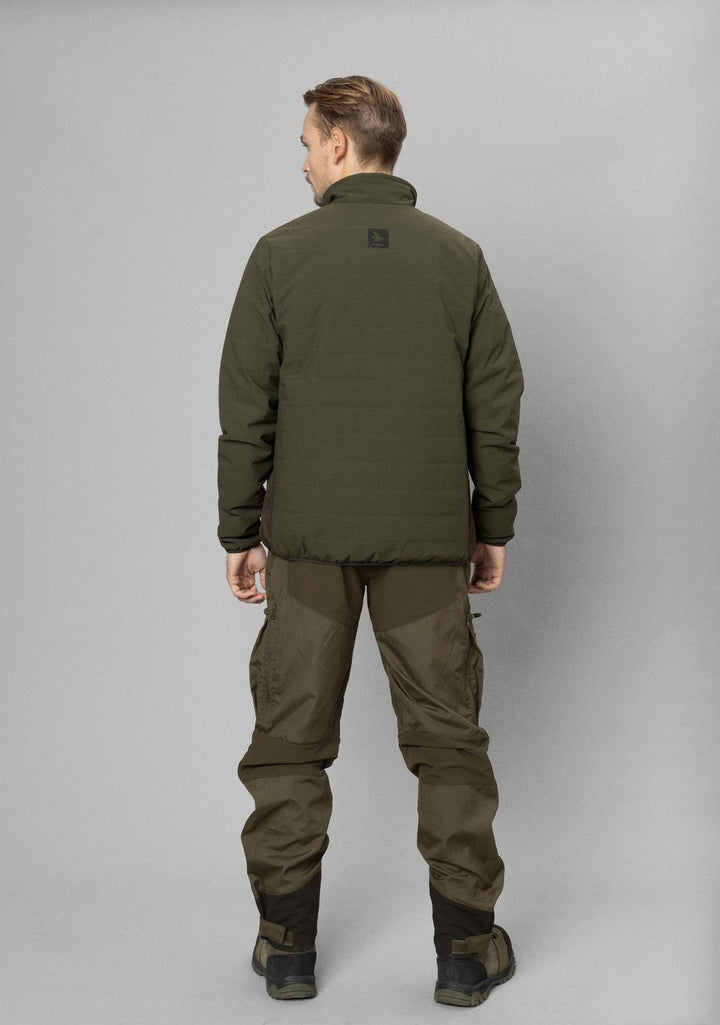 Model photographed from behind in the olive Celsius Heat jacket and matching green trousers against a grey studio wall.