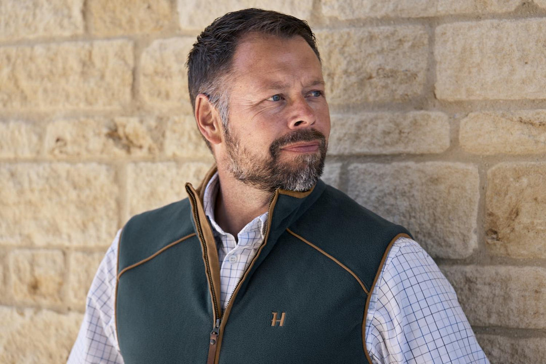 Lifestyle shot: a bearded man in a Härkila Sandhem 200 fleece waistcoat and check shirt stands against a sandstone wall