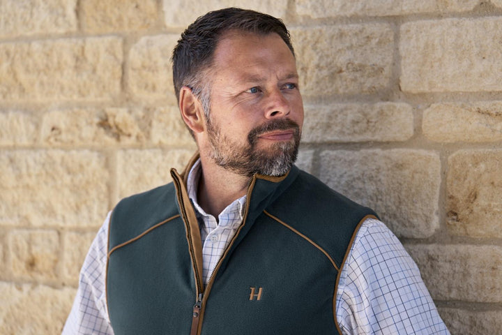 Lifestyle shot: a bearded man in a Härkila Sandhem 200 fleece waistcoat and check shirt stands against a sandstone wall