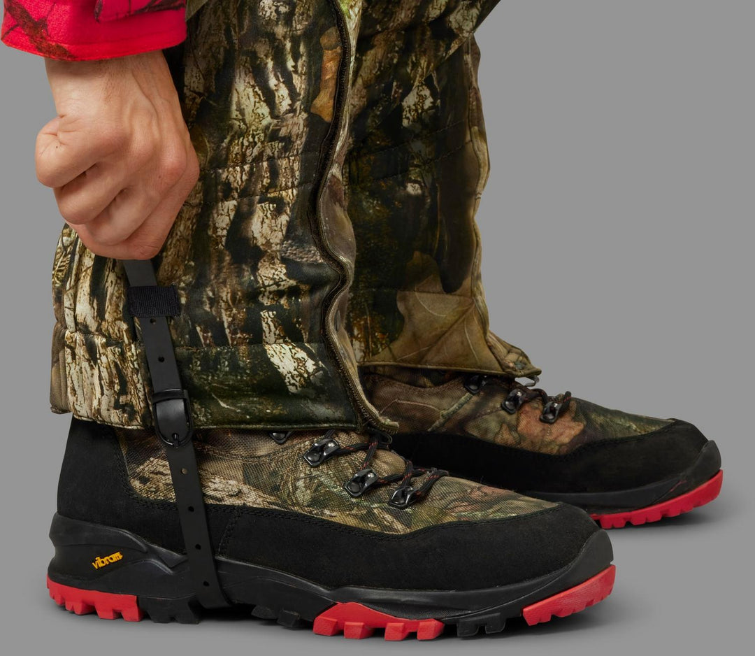 Side close-up of one gaiter fitted over a camo boot, the strap routed under the Vibram sole and buckled at the side.