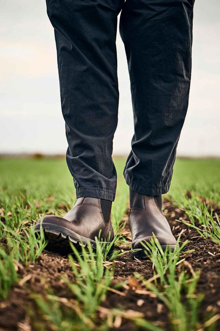 Lifestyle shot of a person in dark trousers walking in brown Grubs Fury S3 safety dealer boots across a green crop field