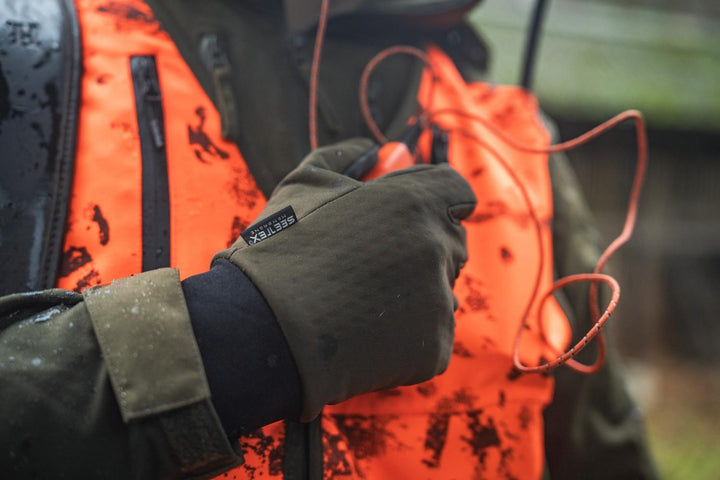 Hunter in a rain-soaked blaze orange and olive jacket with a gloved hand resting on the chest, whistle cord looped across the front.