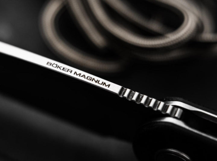Macro close-up of the Böker Magnum engraving on the blade of the Cliff Wharn folding knife with cord in the background