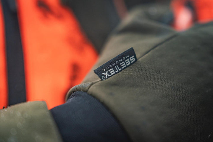 Close-up of the glove cuff against a blaze orange vest, with the Seetex Membrane label stitched onto the pine green softshell.