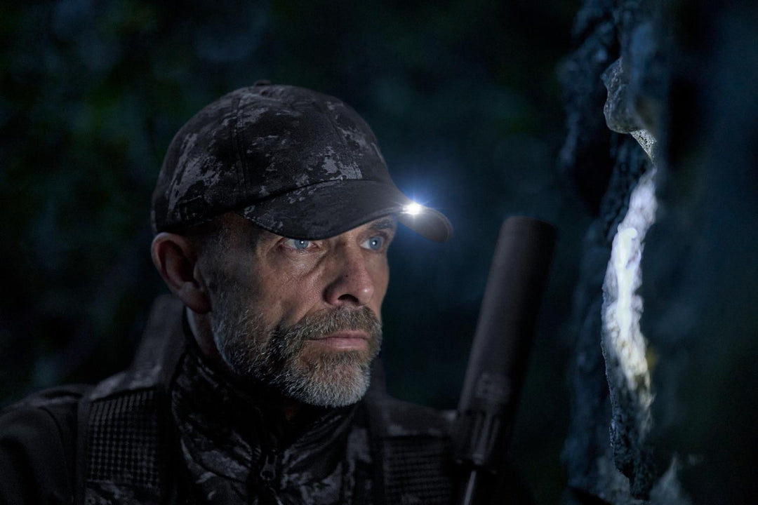 Night lifestyle shot of a hunter wearing the cap with a small head torch clipped to the peak, rifle beside him in the dark.