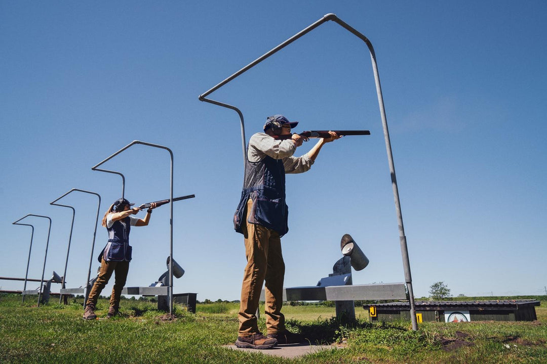Lifestyle: two shooters on a skeet range shooting under high metal gantries, one in a navy Skeet II waistcoat