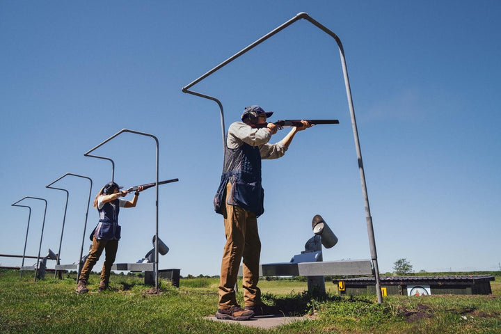 Lifestyle: two shooters on a skeet range shooting under high metal gantries, one in a navy Skeet II waistcoat