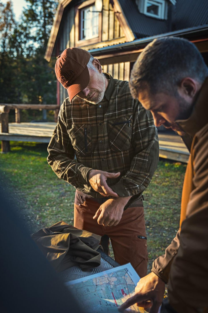 Lifestyle shot: two men outside a timber hunting lodge at dusk, one wearing the Harkila Pajala check shirt, studying a map spread on a tailgate.