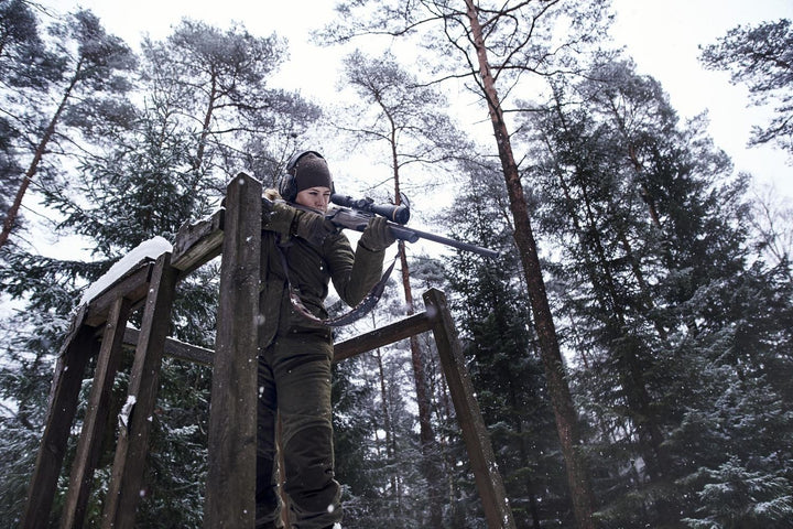 Lifestyle: woman in the olive jacket standing on a wooden high seat in snowy conifer forest, rifle raised