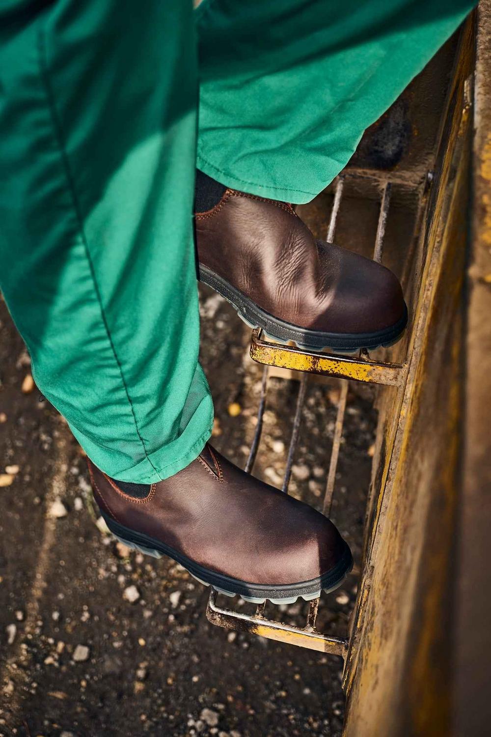 Lifestyle shot of a worker in green overalls wearing brown Grubs Fury S3 safety dealer boots on a wooden farm ladder