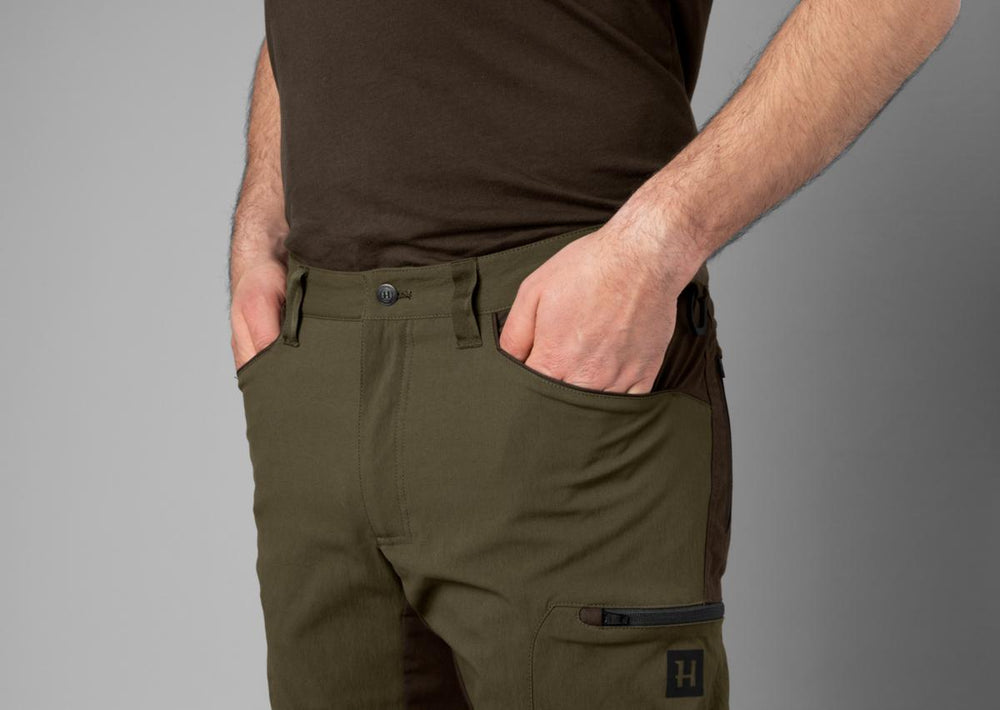 Close-up of a model's left thigh in the willow green Harkila Ragnar trousers, hand in the pocket, showing the zipped leg pocket with Harkila H logo.