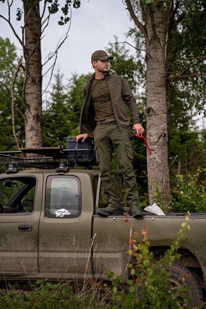 Lifestyle: stalker in Harkila fleece and green trousers standing on the roof rack of a pickup in a birch wood, rifle case beside him