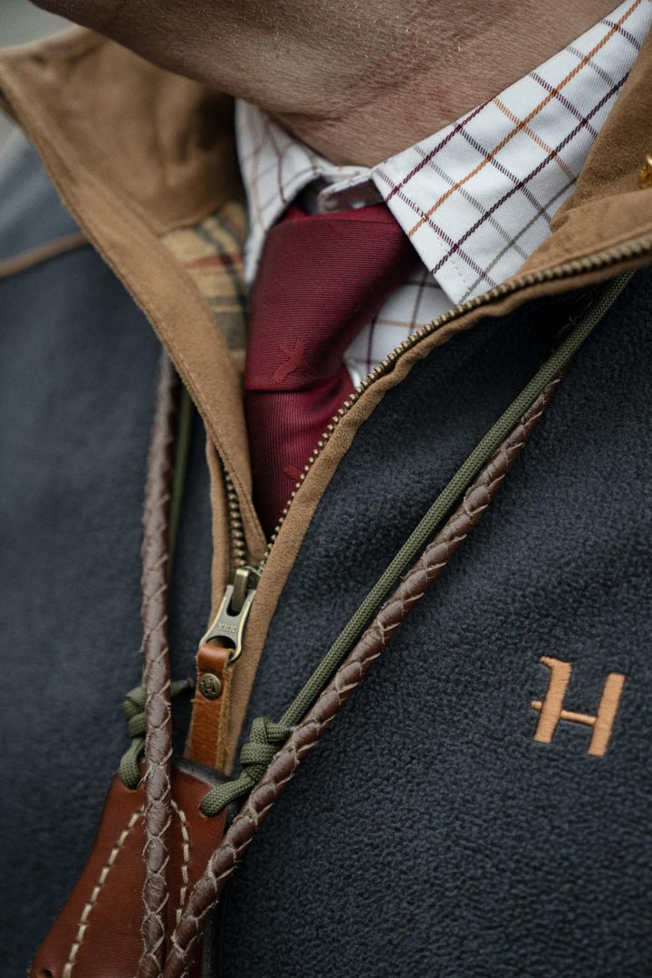 Close-up of the Härkila Sandhem 200 waistcoat zip and copper H logo, worn over a tattersall shirt and burgundy tie with a gun sling across the chest
