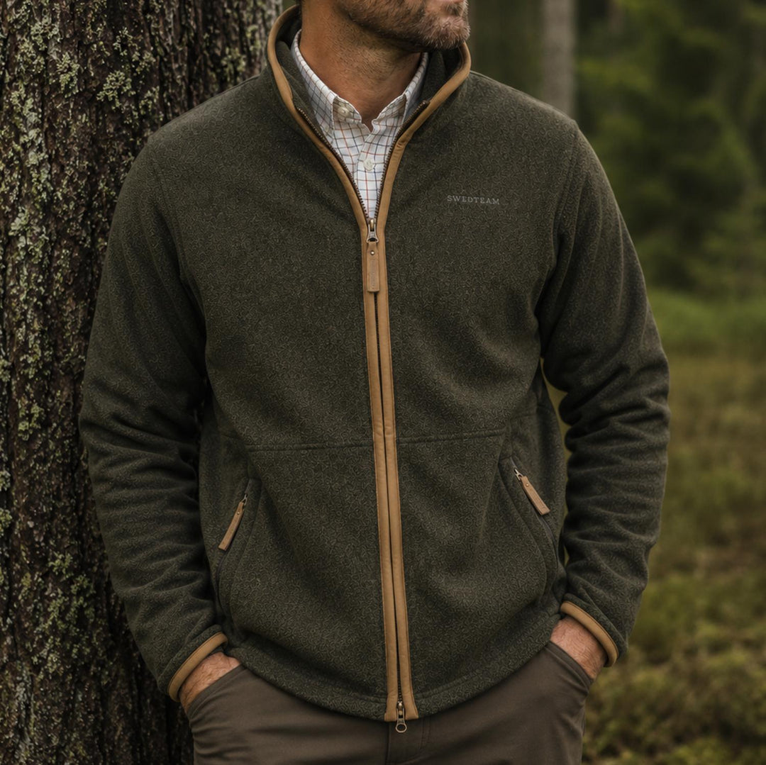 Lifestyle shot: a man leaning against a lichen-covered tree trunk in woodland, wearing the olive fleece unzipped over a tattersall check shirt.