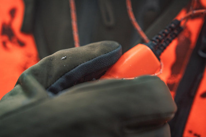 Close-up of a gloved hand gripping an orange object against a blaze orange vest, showing the reinforced thumb and finger panels.