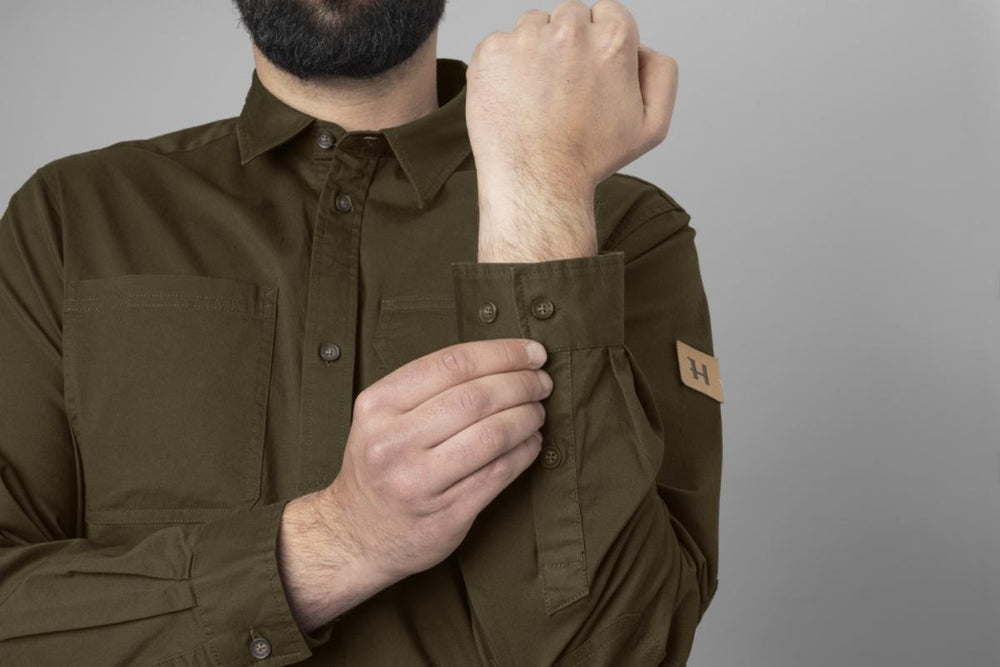 Close-up of a model fastening the buttoned chest pocket flap, with the leather brand badge on the upper sleeve.