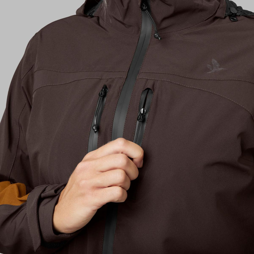 Detail view of a hand drawing up the brown jacket's front zip, showing the taped seams, welded chest pockets and the Seeland bird logo on the chest.